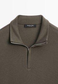 Olivgrüner Reißverschluss-Pullover mit kurzem Stehkragen, aus strukturiertem Material, mit einem silbernen Metallreißverschluss. Markenschild sichtbar.