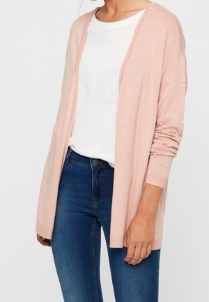 Vrouw draagt een lichtroze open vest met parelversieringen op de mouwen, een wit T-shirt en een blauwe spijkerbroek, gezien van nek tot heupen.