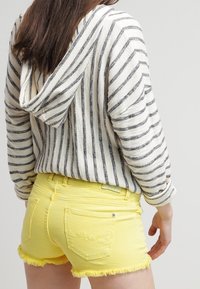 Mujer vestida con una sudadera de rayas blancas y grises y pantalones cortos de mezclilla amarillos brillantes con bordes deshilachados, vista desde atrás.