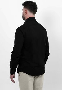 Man met kort donker haar, gekleed in een zwart longsleeve shirt en beige broek, staande met zijn rug naar de camera op een effen witte achtergrond.