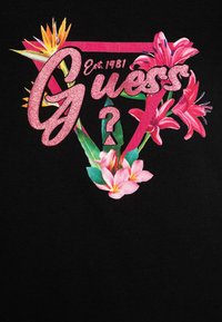 Melns T-krekls ar spīdīgu rozā "Guess" logotipu, rozā liliju ziedu un tropisko lapu florālu dizainu, kā arī dibināšanas gadu "Est. 1981."