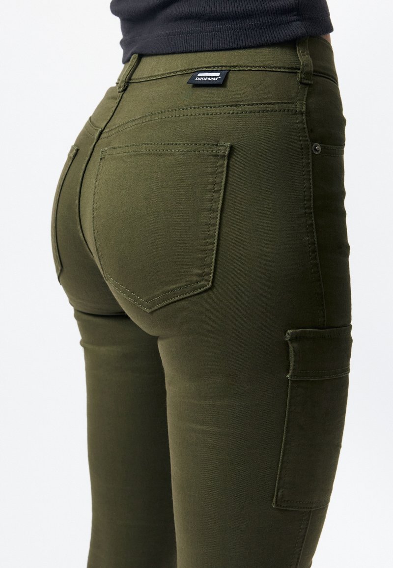 Pantalones cargo de denim elástico en verde oliva, con cinco bolsillos, un detalle en el yugo trasero y una etiqueta de marca cerca de la cintura.