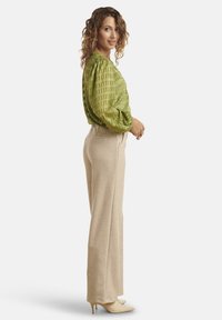 Blusa verde a motivo con maniche a sbuffo, abbinata a pantaloni beige a gamba larga e scarpe con tacco di colore chiaro. Sfondo neutro.