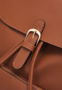 Sac en simili cuir marron avec une fermeture à boucle en métal doré, des accents cousus et un compartiment zippé le long du bord inférieur.