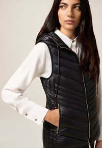 Schwarze gepolsterte Weste mit Kapuze, gestepptem Design, Reißverschluss und Seitentaschen. Über einer weißen Bluse mit geknöpften Manschetten getragen.