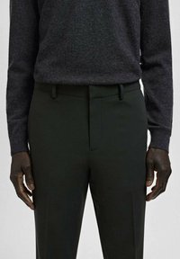 Mężczyzna w ciemnoszarym swetrze z długim rękawem wsuniętym w czarne, dopasowane spodnie, stojący z rękami swobodnie opuszczonymi wzdłuż ciała.