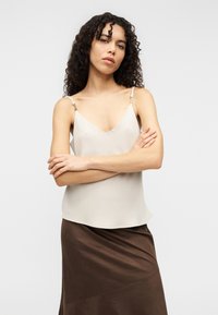 Κρεμ σατινέ καμίσωμα με λεπτές τιράντες και λαιμόκοψη σε σχήμα V, συνδυασμένο με μια σκούρα καφέ, ρέουσα φούστα με διαγώνιες ραφές.