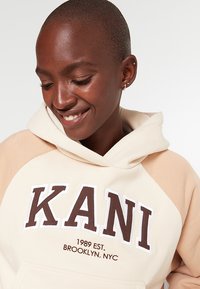 Felpa con cappuccio in crema e beige con logo "KANI" ricamato. Caratterizzata da una texture morbida, tasca a marsupio e design a contrasto sulle spalle.