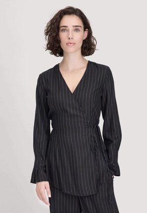Vrouw met kort krullend haar draagt een zwart gestreept wikkelshirt en bijpassende broek, staande voor een effen lichte achtergrond.