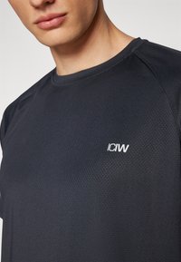 Camiseta atlética oscura hecha de tejido texturizado. Presenta un cuello redondo y un pequeño logo blanco "ICIW" en el pecho.