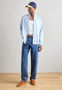 Helles blaues Hemd über einem weißen Tanktop, kombiniert mit lockeren blauen Jeans und braunen Wildleder-Slippers, accessoriert mit einer blauen und hellbraunen Kappe.