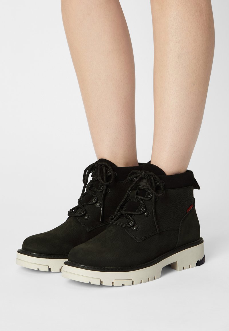 Botas negras hasta el tobillo hechas de cuero texturizado con una suela de goma blanca y lisa. Cuentan con cordones redondos y ojales de metal.