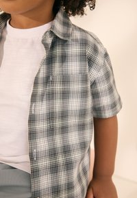 Chemise à manches courtes à carreaux noir et blanc, avec une poche poitrine, une fermeture à boutons, et un tissu léger porté sur un t-shirt blanc.