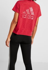 Röd kortärmad träningsskjorta med texturerad yta och en stor glänsande Adidas-logotyp på ryggen, kombinerad med svarta leggings som har vita ränder.