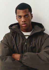 Bruine gewatteerde jas met hood, voorzien van elastische manchetten, een ritssluiting aan de voorkant en trekkoorden. Gedragen over een wit shirt met strepen.