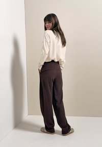 Donna con capelli lunghi e castani, con camicia bianca a maniche lunghe e pantaloni marrone scuro, in piedi con le mani nelle tasche, che guarda indietro oltre la spalla.