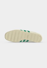 Σόλα από μπεζ αθλητικό παπούτσι Adidas με πράσινες λωρίδες, με σχέδιο ψαροκόκαλο και το λογότυπο Adidas κοντά στην καμάρα.