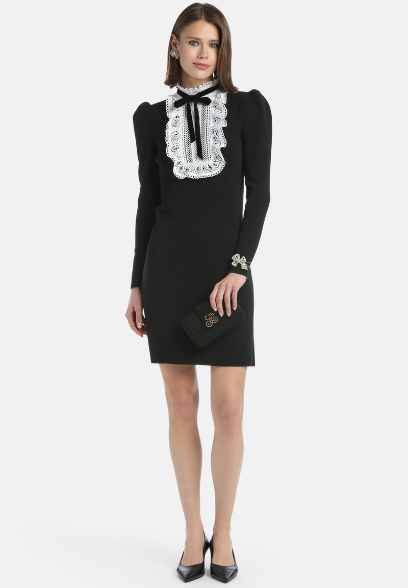 Schwarzes Kleid mit langen Ärmeln, weißem Spitzenkragen und Schleifendetail. Inklusive schwarzer Clutch und silber-aczentuierten Manschetten. Schwarze Schuhe mit Absatz.