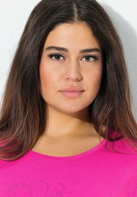 Světlý růžový top s krátkým rukávem a kulatým výstřihem. Modelka má dlouhé hnědé vlasy, hladkou pleť a nosí jemný make-up. Jednoduché pozadí.