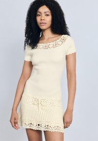 Φόρεμα με ριμπ πλέξη σε κρεμ χρώμα, με λουλουδάτο κέντημα στον λαιμό και στο τελείωμα. Διαθέτει κοντά μανίκια, ζώνη στη μέση και υφασμένο κεντητό τελείωμα με υφή.