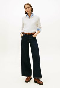 Pull en maille texturée blanche, chemise bleue à col en dessous, jeans noirs à jambes larges, ceinture en cuir marron, et mocassins en cuir marron à semelles épaisses.