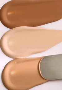 Flytende foundation swatcher i tre nyanser: mellombrun, beige og lys krem, med en jevn tekstur påført med en spatel på en hvit overflate.