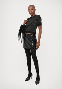 Top corto in maglia nera con motivo logo, mini gonna in vinile nero lucido, collant neri opachi e décolleté nere con punta. Borsa nera con frange.