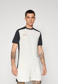 Koszulka sportowa Nike z krótkim rękawem, z jasnoszarym korpusem, czarnymi rękawami, czerwonym logo i akcentami po bokach. Materiał wydaje się gładki i dopasowany.