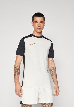 Nike trumpas rankovėmis sportinis marškinėliai, turintys šviesiai pilką kūną, juodas rankoves, raudoną logotipą ir šonines akcentus. Audinys atrodo glotnus ir prigludęs.