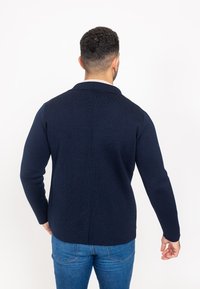 Merine sinine kootud kardigan tekstuuritud viimistlusega, millel on nugaga krae ja pikad varrukad, kantud džinside peal.