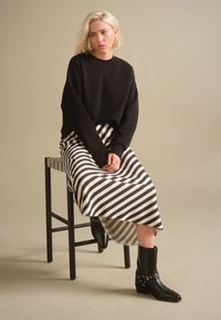 Blondynka w czarnym swetrze, czarno-białej pasiastce spódnicy i czarnych botkach siedzi na drewnianym stołku na tle jednolitego beżowego tła.