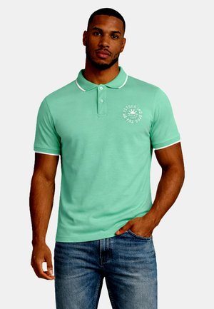 Man met baard die een mintgroen poloshirt met witte bies en tekst op de borst draagt, blauwe spijkerbroek, staand met één hand in de zak.