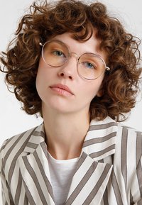 Lunettes rondes en métal avec des verres teintés ambre et de fines montures en argent. Le modèle porte un blazer rayé sur une chemise blanche, avec des cheveux bouclés.
