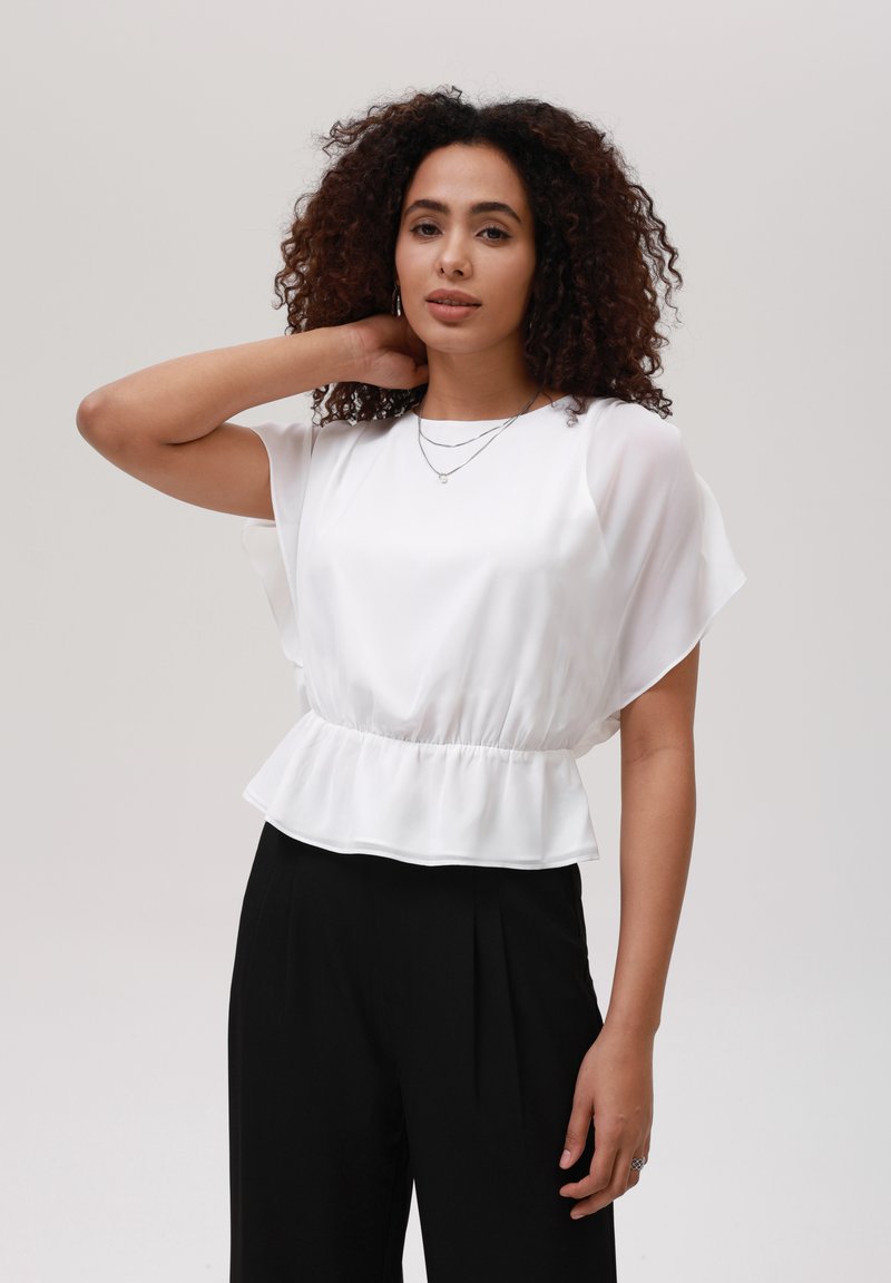 Top peplum bianco con maniche corte e drappeggiate e vita elastica, abbinato a pantaloni neri. Texture liscia, design minimalista.