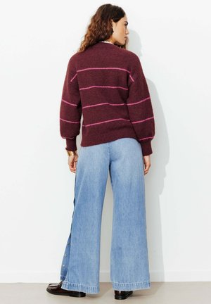 Femme aux cheveux ondulés portant un pull marron avec des rayures roses, un jean large bleu clair et des chaussures foncées, debout contre un mur blanc.