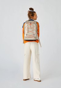 Beige Rucksack mit orangen Akzenten, ausgestattet mit verstellbaren Trägern, einem Kordelzugverschluss und einem schlanken, modernen Design.