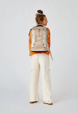 Beige Rucksack mit orangen Akzenten, ausgestattet mit verstellbaren Trägern, einem Kordelzugverschluss und einem schlanken, modernen Design.