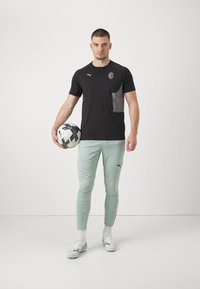 Camiseta deportiva negra con panel gris, logo en el pecho; leggings verde menta claros; botas de fútbol blancas; sosteniendo un balón de fútbol en blanco y negro.