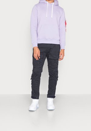 Homme portant un sweat à capuche lavande, un pantalon cargo noir avec plusieurs poches, et des baskets montantes blanches, debout devant un fond clair uni.