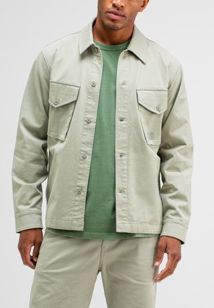 Hellgrüne Jacke mit zwei vorderen Taschen, Knopfverschluss und Kragen, getragen über einem dunkelgrünen Shirt, kombiniert mit passenden Hosen.