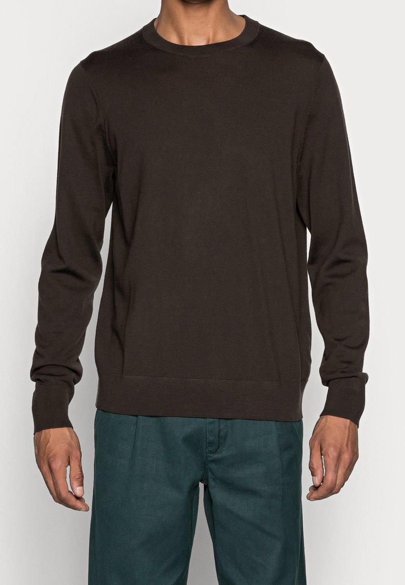 Homme portant un pull marron foncé à manches longues avec col rond et un pantalon bleu sarcelle, debout devant un fond clair uni.