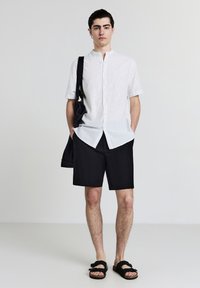 Junger Mann steht mit den Händen in den Taschen, trägt ein weiß gestreiftes Kurzarmhemd, schwarze Shorts, schwarze Sandalen und eine schwarze Umhängetasche.