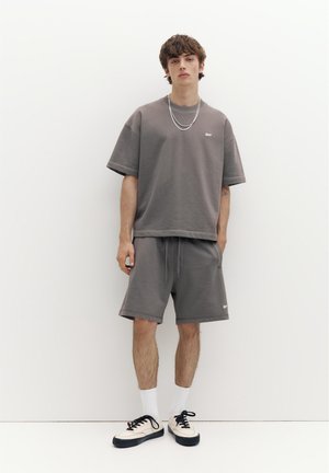 Młody mężczyzna w pasującym szarym, luźnym t-shircie i krótkich spodenkach, białych skarpetkach, czarno-białych tenisówkach oraz z nałożonymi warstwowo srebrnymi łańcuszkami, stojący przy białej ścianie.