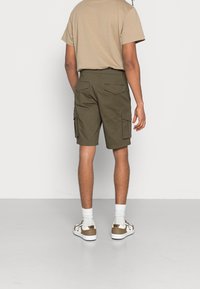 Pantaloni cargo verde oliva con multiple tasche laterali, abbinati a una maglietta beige e calzini bianchi. Le calzature presentano accenti marroni.