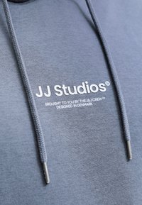 Sweat à capuche gris avec texte blanc "JJ Studios" et un texte plus petit indiquant un design par The J&J Crew au Danemark, avec des cordons gris à embouts métalliques.