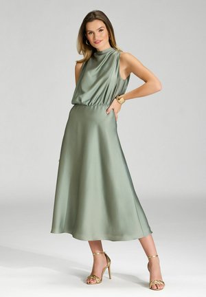 Vestido verde satinado sin mangas con un escote alto, cintura fruncida y falda en línea A que fluye. Combinado con sandalias de tacón dorado metálico.