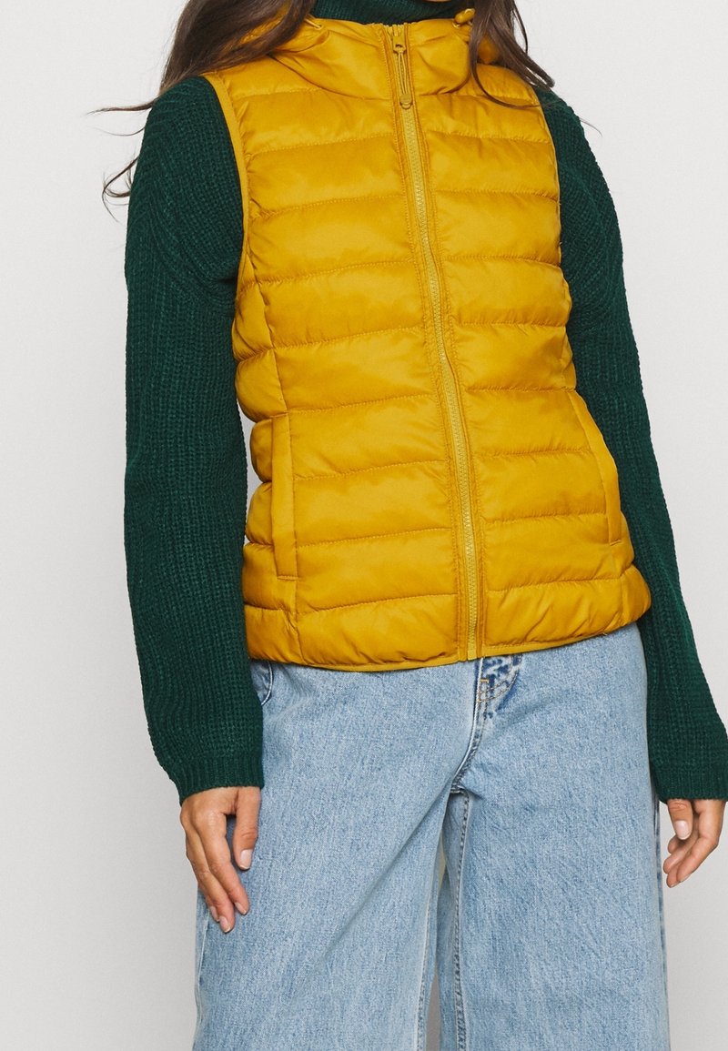 Gilet imbottito giallo con colletto alto, chiusura a zip e texture trapuntata, abbinato a un maglione lavorato a maglia verde e jeans in denim blu chiaro.