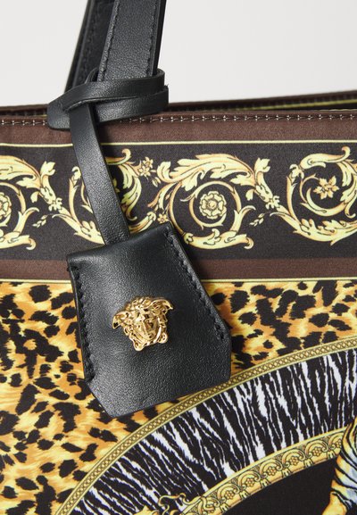Prim-plan al unei genți de tip tote cu mânere din piele neagră, emblemă ornate cu capul de Medusa de culoare aurie și material cu imprimeu de animal în nuanțe de brun, negru și auriu.