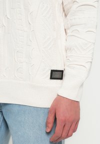 Close-up van de hand van een persoon die naast hun lichaam rust, gekleed in een witte gestructureerde gebreide trui en lichte blauwe denim jeans.
