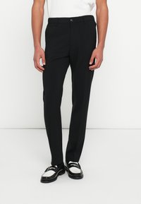 Zwarte op maat gemaakte broek met een rechte pijp, voorzien van een tailleband met knopen en zakken. Gecombineerd met zwart-witte loafers.